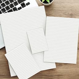 A collection of lined note pads in various sizes, featuring a light colour scheme, on a wooden desk beside a laptop.